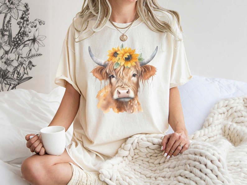 May include: A cream colored t-shirt with a watercolor illustration of a brown and white cow wearing a sunflower crown.