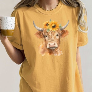 May include: A yellow t-shirt with a watercolor illustration of a brown Highland cow wearing a sunflower crown.