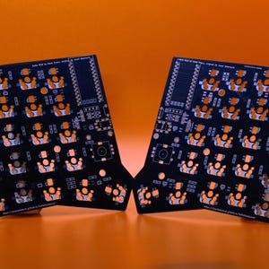 May include: Two black printed circuit boards for a split keyboard, featuring an orange solder mask pattern with numerous switch footprints, mounting holes, and small white text labels printed across the surface of each board.
