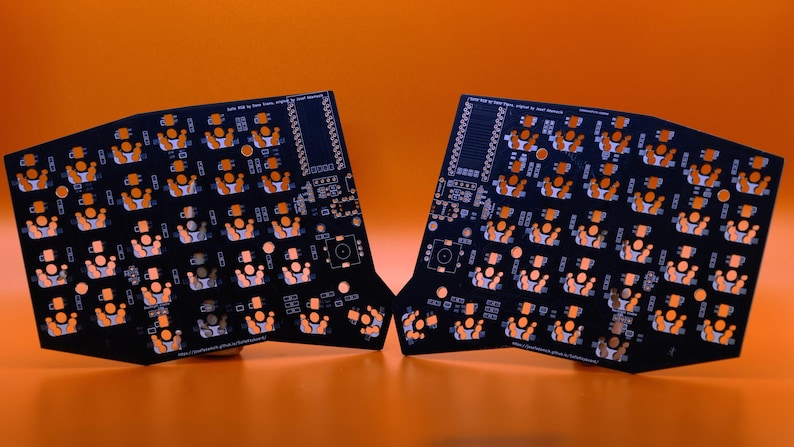 May include: Two black printed circuit boards for a split keyboard, featuring an orange solder mask pattern with numerous switch footprints, mounting holes, and small white text labels printed across the surface of each board.