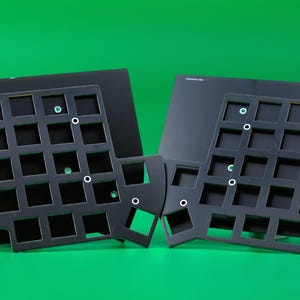 May include: Two black, split-style mechanical keyboard plates featuring a grid of square cutouts for switches. Each plate has a distinct, ergonomic, non-rectangular shape with several circular mounting holes distributed across the frame.