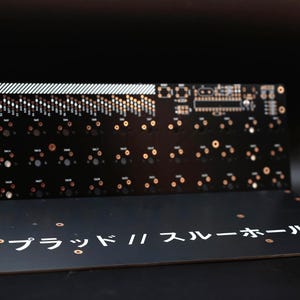 May include: A black printed circuit board for a keyboard with copper-colored contact points and white markings. The board features a grid of switch sockets and the Japanese text "プラッド // スルーホール" printed in white on the lower section.