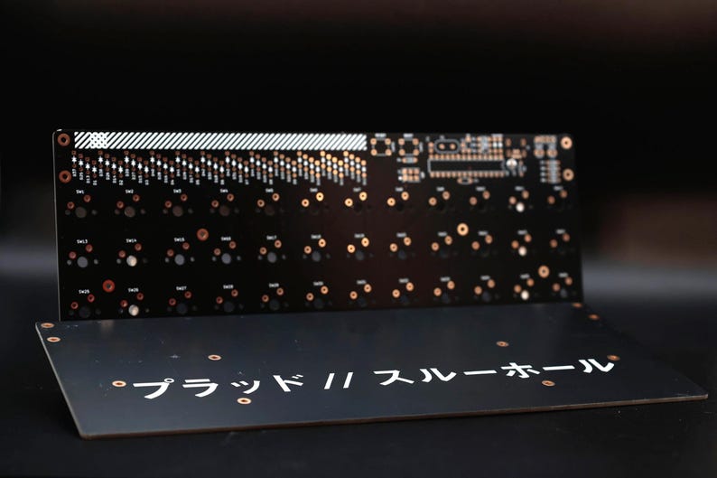 May include: A black printed circuit board for a keyboard with copper-colored contact points and white markings. The board features a grid of switch sockets and the Japanese text "プラッド // スルーホール" printed in white on the lower section.