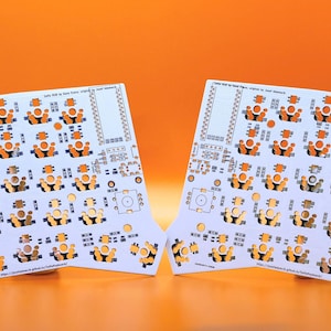 May include: Two white printed circuit boards for a split keyboard, featuring a symmetrical layout with numerous circular orange switch sockets, various electronic component footprints, and small text labels printed across the surface.