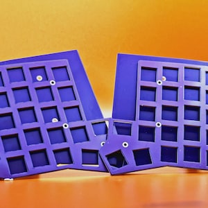 May include: A pair of purple split keyboard plates featuring a grid of square cutouts for mechanical switches. Each plate has an irregular, angled shape with several small circular mounting holes distributed across the surface for assembly.