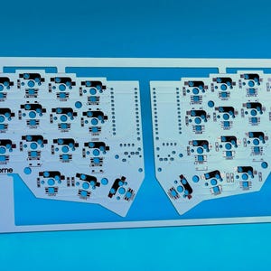 May include: A white printed circuit board for a split mechanical keyboard, featuring two symmetrical halves connected by a top frame. The board includes numerous switch sockets and LED pads, with the text "corne" printed on each section.