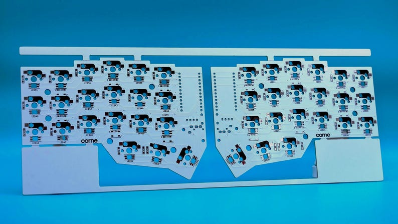 May include: A white printed circuit board for a split mechanical keyboard, featuring two symmetrical halves connected by a top frame. The board includes numerous switch sockets and LED pads, with the text "corne" printed on each section.
