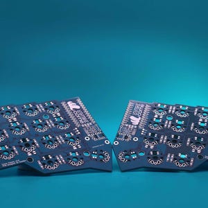 May include: Two dark blue printed circuit boards for a split mechanical keyboard. Each board features a grid of circular switch sockets, white printed labels, and the text "crkbd" with a logo. The boards have a distinct ergonomic shape.