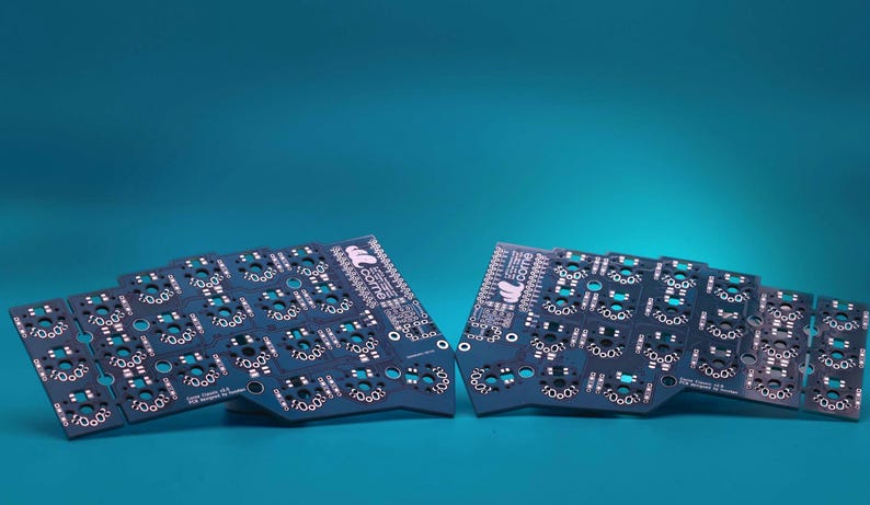May include: Two dark blue printed circuit boards for a split mechanical keyboard. Each board features a grid of circular switch sockets, white printed labels, and the text "crkbd" with a logo. The boards have a distinct ergonomic shape.