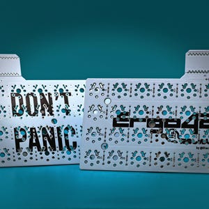 May include: Two white printed circuit boards for a mechanical keyboard. The left board features the black text "DON'T PANIC" and the right board features the black text "Ergo42" with "by @Fiacco42" printed below it. Both boards have numerous holes.