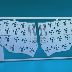 May include: A white printed circuit board for a split mechanical keyboard, featuring a symmetrical layout with numerous circular switch cutouts and mounting holes. The board includes the text "Corne" and "Corne Cherry V3.0 PCB designed by foostan".