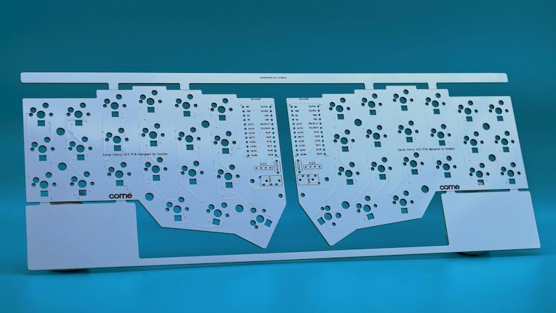 May include: A white printed circuit board for a split mechanical keyboard, featuring a symmetrical layout with numerous circular switch cutouts and mounting holes. The board includes the text "Corne" and "Corne Cherry V3.0 PCB designed by foostan".