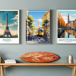 May include: Three framed travel art prints featuring iconic cityscapes: Paris, Lisbon, and Amsterdam. The Paris print shows the Eiffel Tower, the Lisbon print features a yellow tram, and the Amsterdam print depicts canals. A wooden surfboard rests on a wooden shelf below.