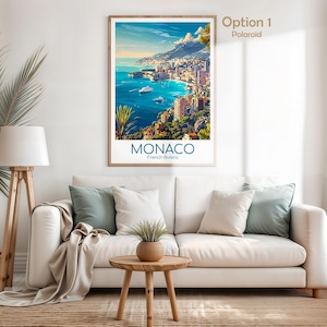 Puede incluir: Un p&oacute;ster enmarcado de M&oacute;naco, Riviera francesa, con una vista de una ciudad costera en tonos azules, verdes y marrones. El p&oacute;ster se exhibe sobre un sof&aacute; blanco con cojines, una mesa auxiliar de madera y una l&aacute;mpara de pie.