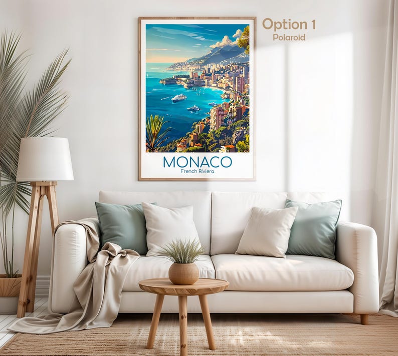 Puede incluir: Un p&oacute;ster enmarcado de M&oacute;naco, Riviera francesa, con una vista de una ciudad costera en tonos azules, verdes y marrones. El p&oacute;ster se exhibe sobre un sof&aacute; blanco con cojines, una mesa auxiliar de madera y una l&aacute;mpara de pie.