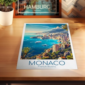 Puede incluir: Una impresi&oacute;n de M&oacute;naco, Riviera Francesa, que muestra una ciudad costera con edificios, barcos y un mar azul bajo un cielo brillante. La palabra "MONACO" est&aacute; impresa en azul en la parte inferior de la impresi&oacute;n.