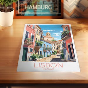 May include: A rectangular art print featuring a colorful illustration of a narrow street in Lisbon, Portugal. The scene depicts pastel-colored buildings with balconies, green plants, and a stone path under a blue sky. The text LISBON Portugal is printed below.