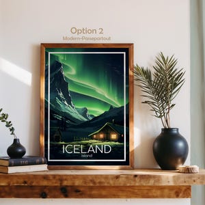 May include: A framed print of Iceland's landscape, featuring the Northern Lights. The artwork showcases a cabin with lit windows, mountains, and the text "ICELAND Island". The frame is a warm brown color.