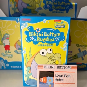 Peut inclure: Présentation de boîtes de collection Bikini Bottom Buddies avec des personnages de poissons de dessins animés. Les boîtes sont jaunes et bleues avec le logo Bob l'éponge. Une carte d'identité rose avec "Limp Fish" est devant.