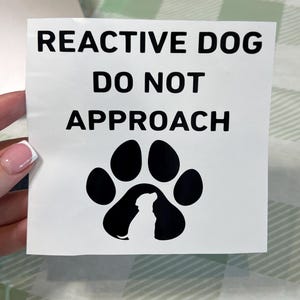 Peut inclure: Autocollant carré blanc avec texte noir : "REACTIVE DOG DO NOT APPROACH". En dessous, une empreinte de patte noire avec la silhouette blanche d'un chien. L'autocollant est tenu par une main.