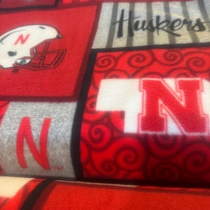 May include: Red, white, and gray fleece fabric featuring the word "NEBRASKA" in white block letters, the word "Huskers" in black script, and the letter "N" in red. The fabric also displays a football helmet and a cartoon figure.