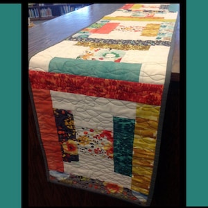 May include: A colorful quilted table runner with the text "Boxed In." The runner features a patchwork design with various fabrics in shades of red, orange, teal, yellow, and floral patterns. The design is by Tricia Lynn Maloney for Villa Rosa Designs.