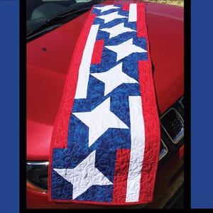 May include: A patriotic quilt with a red, white, and blue design featuring white stars on a blue background, and red and white stripes. The text "BANNER DAY" is at the top. The quilt is displayed on a red car bonnet.