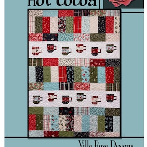 May include: A patchwork quilt with the words "Hot Cocoa" at the top. The quilt features squares of various fabrics in red, green, and white, with rows of cocoa cup designs. The bottom of the quilt has the words "Villa Rosa Designs".