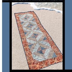 May include: A rectangular quilt with a beach theme, featuring a design of triangles and starfishes in shades of blue, tan, and orange. The quilt is set on a sandy beach with the text "Beachcomber" at the top. The quilt is designed by Tricia Lynn Maloney.