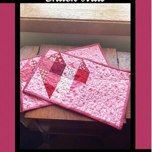 May include: A rectangular quilted snack mat featuring a patchwork heart design in shades of pink and red. The fabric displays various floral and geometric patterns with intricate stitching. The text Dear Heart Snack Mat, Tricia Lynn Maloney, Orphan Quilt Designs, and for Villa Rosa Designs is printed on the border.