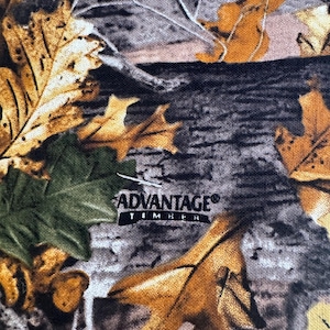 Pode incluir: Tecido camuflado com um padrão de folhas castanhas e amarelas, folhas verdes e casca de árvore cinzenta. O texto "ADVANTAGE TIMBER" está impresso no tecido.