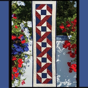 May include: A quilted table runner with a patriotic theme. The runner features a repeating pattern of red, white, and blue stars and ribbons. The text "Stars & Ribbons" is at the top. The design is by Tricia Lynn Maloney.