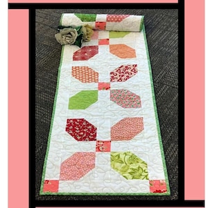 May include: A quilted table runner with a floral pattern in shades of red, pink, green, and orange. The runner has a white background and is bordered with green. The text "Cotton Candy" is at the top.
