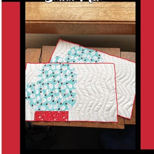 May include: Two quilted snack mats with a winter theme. One mat features a light blue snowman pattern, and the other has a white background with wavy stitching. The mats are trimmed with red binding. The text "Woolly Mitten Snack Mat" is at the top.