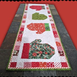 May include: A quilted table runner featuring mitten designs in red, green, and patterned fabrics. The words "COLOR MITTENS" are at the top. The bottom text reads "Sewl Sister for Villa Rosa Designs". The runner is on a dark gray surface.