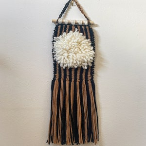 Puede incluir: Un tapiz de pared de macramé hecho a mano con un pompón central blanco crema. El diseño incorpora hilo negro y marrón, con una varilla de madera para colgar. La parte inferior tiene largas borlas con flecos.