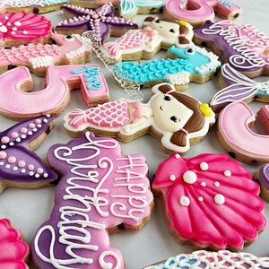 May include: A collection of decorated sugar cookies in various shapes, including starfish, seashells, mermaid tails, and seahorses. The cookies are primarily pink, purple, and blue, with white icing details. Some cookies have the number "5" and the words "Happy Birthday".