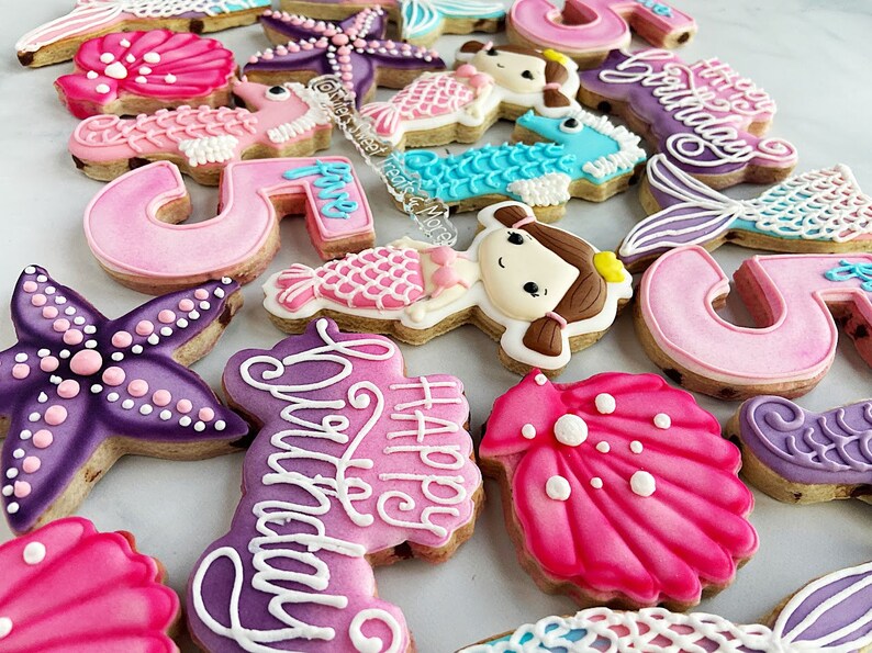 May include: A collection of decorated sugar cookies in various shapes, including starfish, seashells, mermaid tails, and seahorses. The cookies are primarily pink, purple, and blue, with white icing details. Some cookies have the number "5" and the words "Happy Birthday".