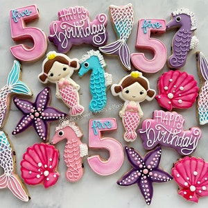 May include: A collection of decorated sugar cookies in a mermaid theme. The cookies include mermaid tails, starfish, seahorses, mermaids, the number "5", and the words "Happy Birthday". The cookies are decorated with pink, purple, blue, and white icing.