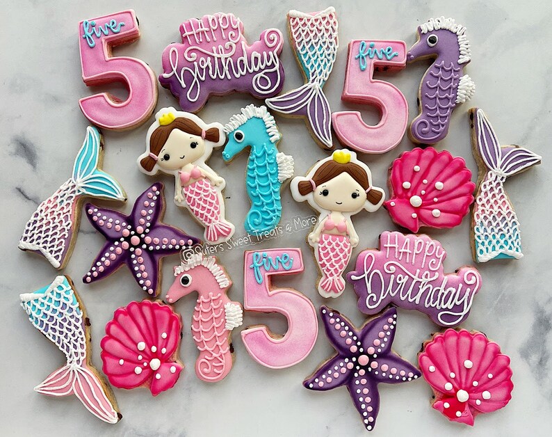 May include: A collection of decorated sugar cookies in a mermaid theme. The cookies include mermaid tails, starfish, seahorses, mermaids, the number "5", and the words "Happy Birthday". The cookies are decorated with pink, purple, blue, and white icing.