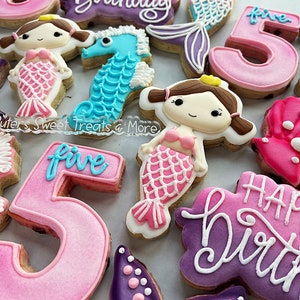 May include: A collection of decorated sugar cookies in a mermaid theme. Cookies include mermaid figures, seahorses, and the number "5" in pink and blue. Some cookies have the words "Happy Birthday" written in white icing.