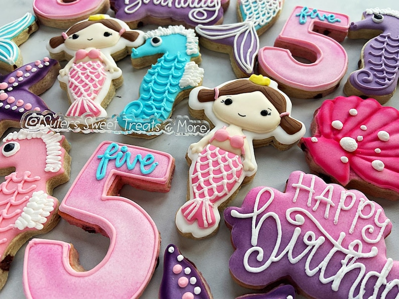 May include: A collection of decorated sugar cookies in a mermaid theme. Cookies include mermaid figures, seahorses, and the number "5" in pink and blue. Some cookies have the words "Happy Birthday" written in white icing.