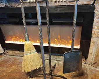 Könnte beinhalten: Ein Schmiedeeisernes Kaminbesteck mit Ständer. Das Set beinhaltet einen Besen, einen Schürhaken, eine Schaufel und ein Design mit gedrehtem Griff. Die Werkzeuge werden vor einem Kamin mit Steinfassung präsentiert.