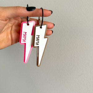 Könnte beinhalten: Zwei Schlüsselanhänger in Blitzform mit dem Wort "MAMA" darauf. Ein Schlüsselanhänger ist pink und weiß, der andere ist braun und weiß.