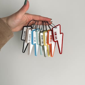 May include: A set of ten colorful lightning bolt-shaped keychains with different names printed on each. The keychains are arranged in a rainbow pattern, with the red keychain at the top and the black keychain at the bottom.