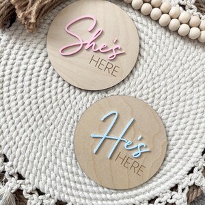 Puede incluir: Dos letreros redondos de madera con las frases "She's HERE" en rosa y "He's HERE" en azul. Se encuentran sobre una superficie tejida de color blanco roto, con cuentas de madera y detalles de cuerda. Un anuncio encantador.