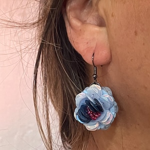 May include: A pair of dangle earrings featuring a blue and iridescent flower design. The flower is made of sequins and has a pink centre.