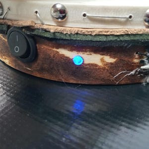 May include: Close-up of a handcrafted object with a brown, textured surface and a white top. It features a black on/off switch, silver studs, and a blue light. The object has a rustic, artisanal aesthetic.