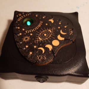 May include: A black plastic base with a power switch supports a dark wooden planchette featuring engraved moon phases, an eye, stars, and the words HELLO, GOODBYE, YES, and NO. A small circular cutout reveals a glowing green LED light.