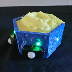 May include: A blue hexagonal box with a detailed dragon sculpture inside. The box has a swirling pattern on the sides and is illuminated with green lights. Two black sensors with white spheres are attached to the sides.